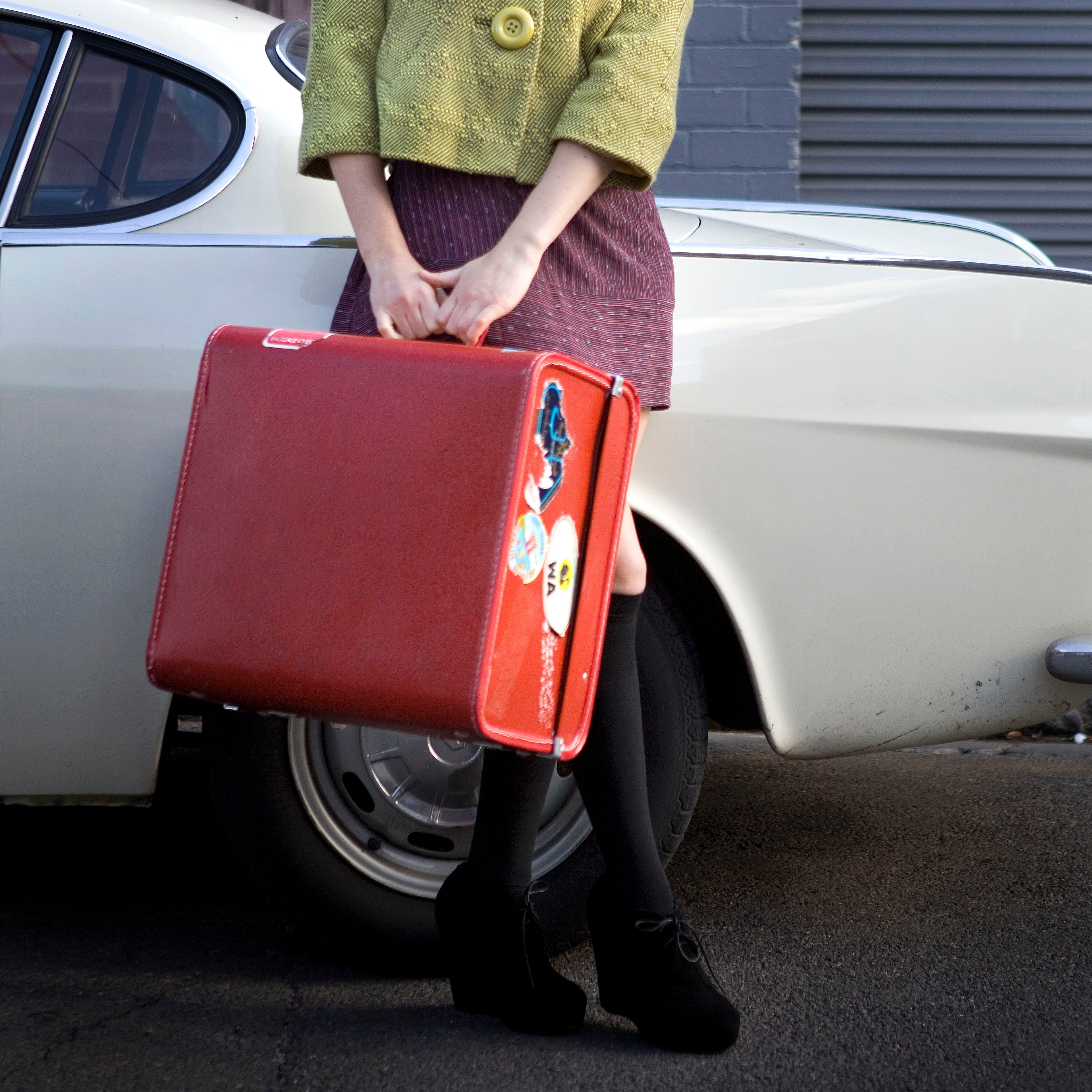 Person stands by a vintage car, holding a suitcase. They wear a textured jacket and skirt, with a poised, classic style