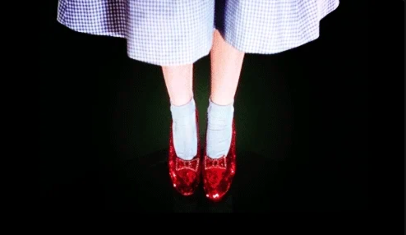 A person wearing sequined shoes and socks, with a dress featuring a checkered pattern, stands on a dark background