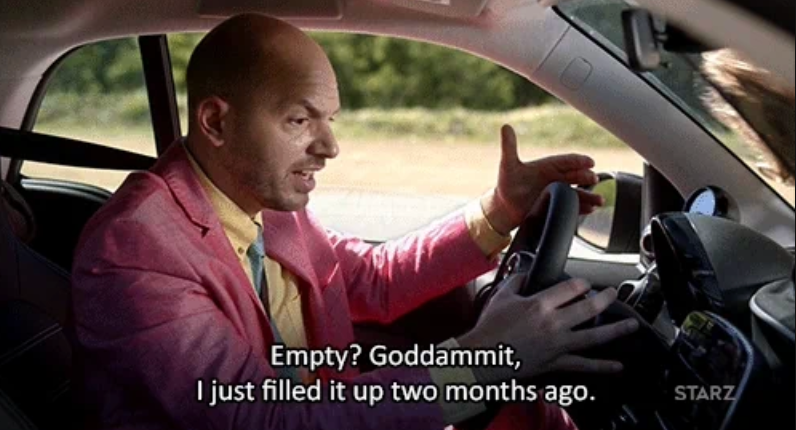A man in a car, wearing a suit, expresses frustration about an empty gas tank, saying he filled it two months ago