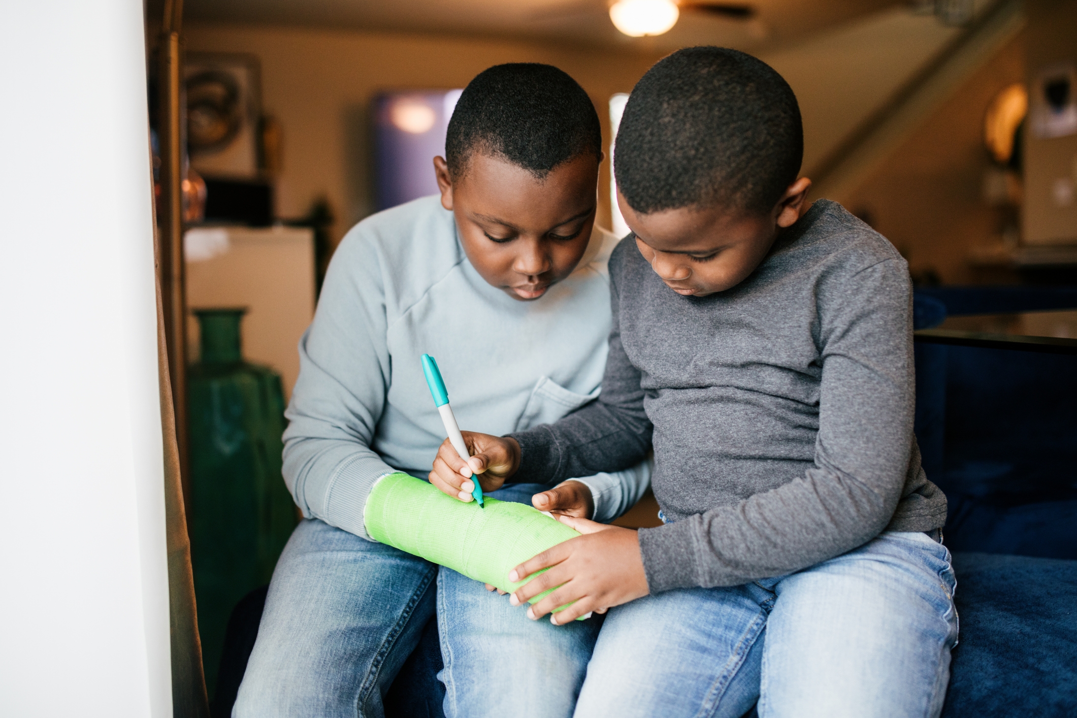 Two children sitting together; one is writing on the other's cast with a marker
