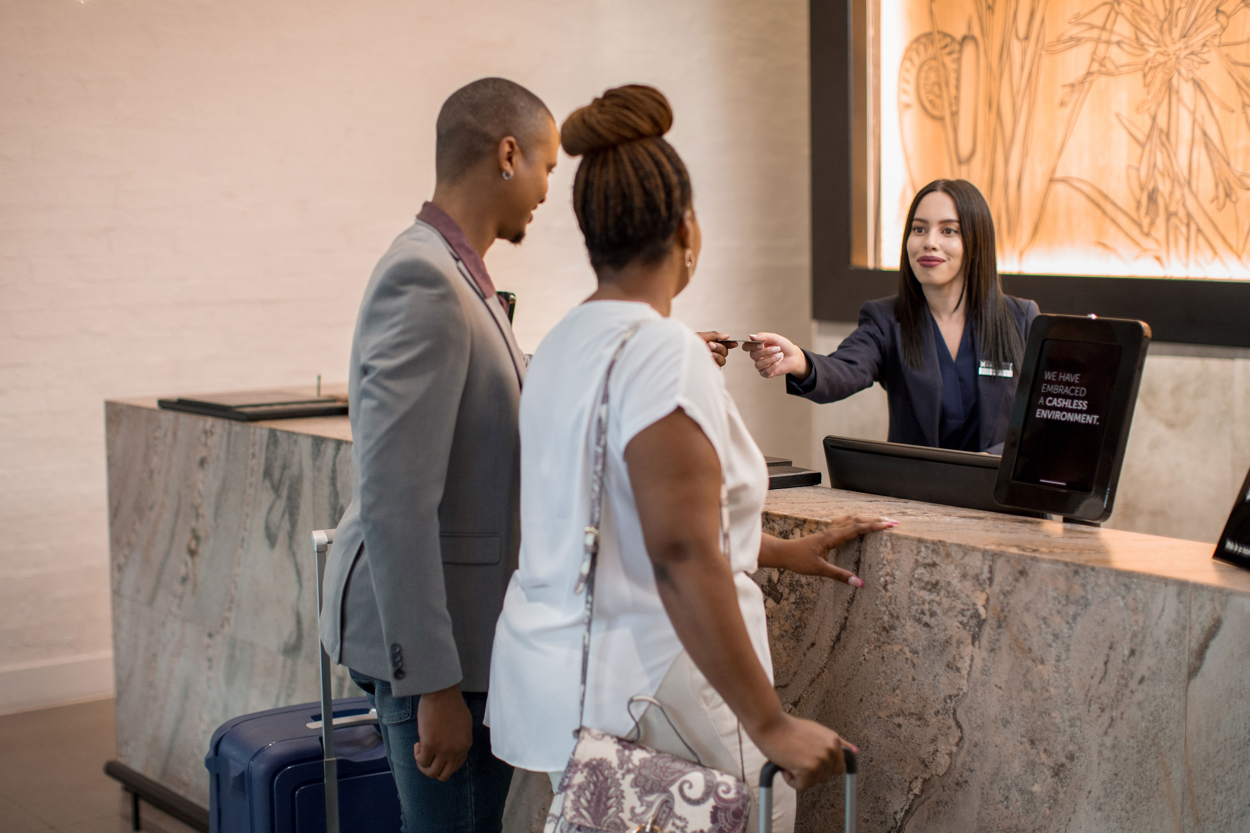 Hotel check-in scene with a smiling receptionist handing a key card to a couple at the front desk. Suitcase visible next to them