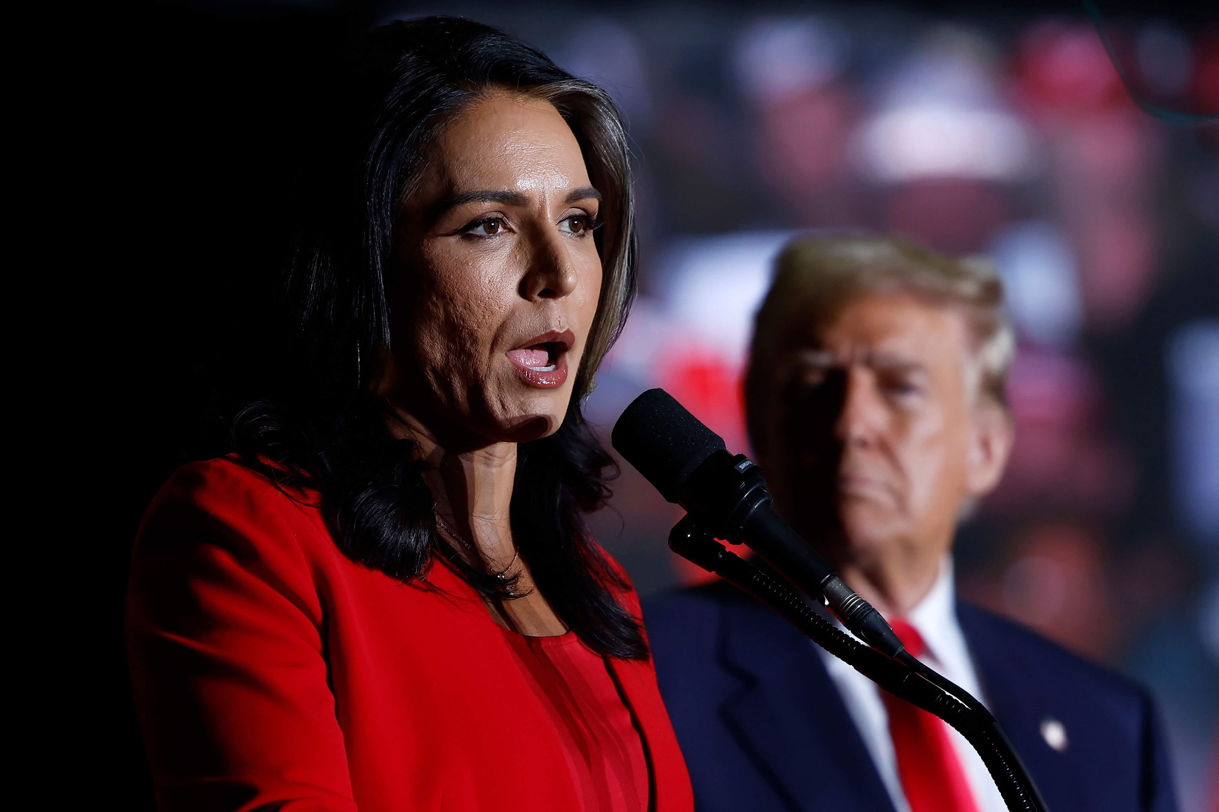 Two people are speaking at an event. The person in focus is wearing a formal suit and is speaking into a microphone. Another person is in the background