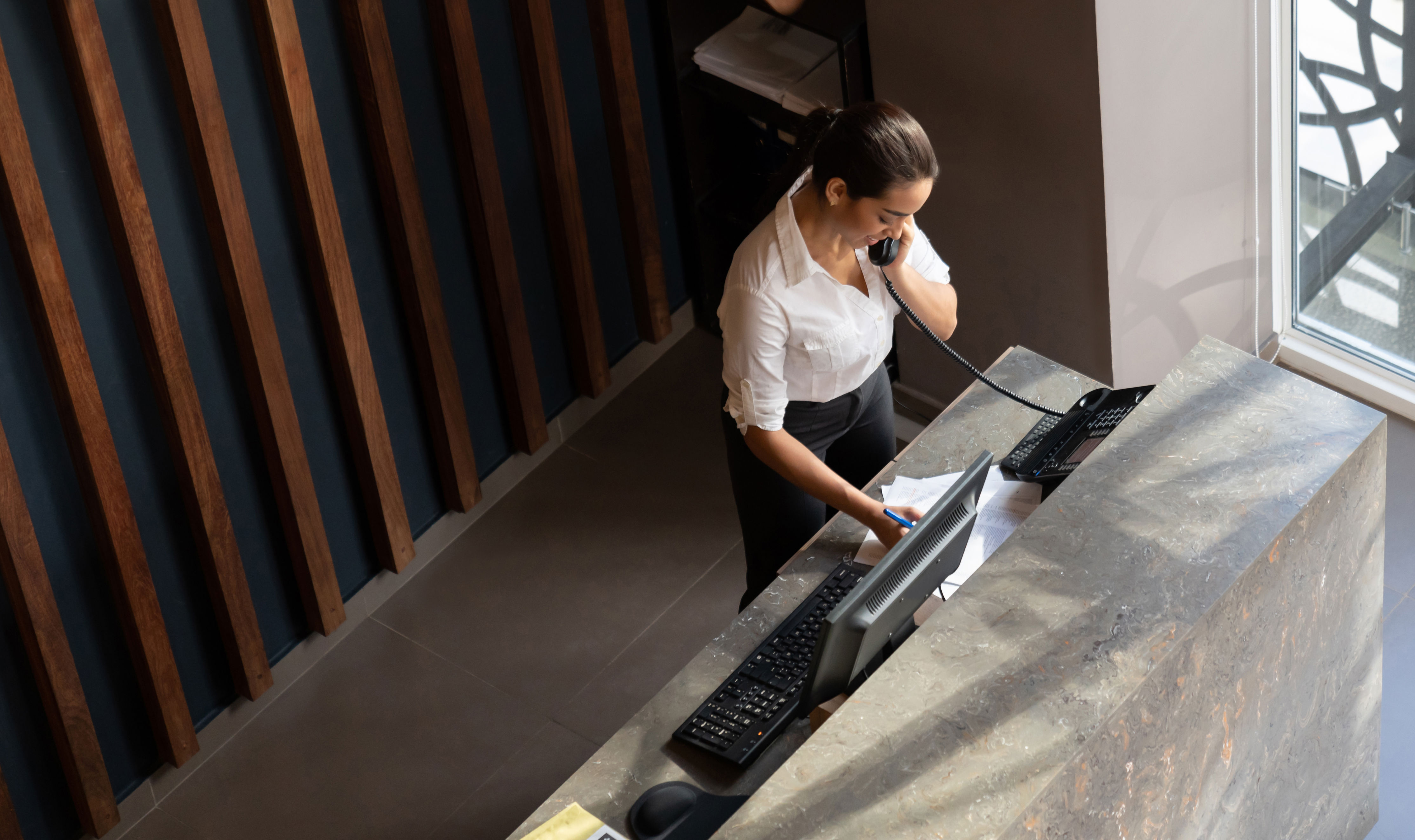 A hotel receptionist at a desk speaks on the phone while checking documents, with a computer and keyboard in front of her