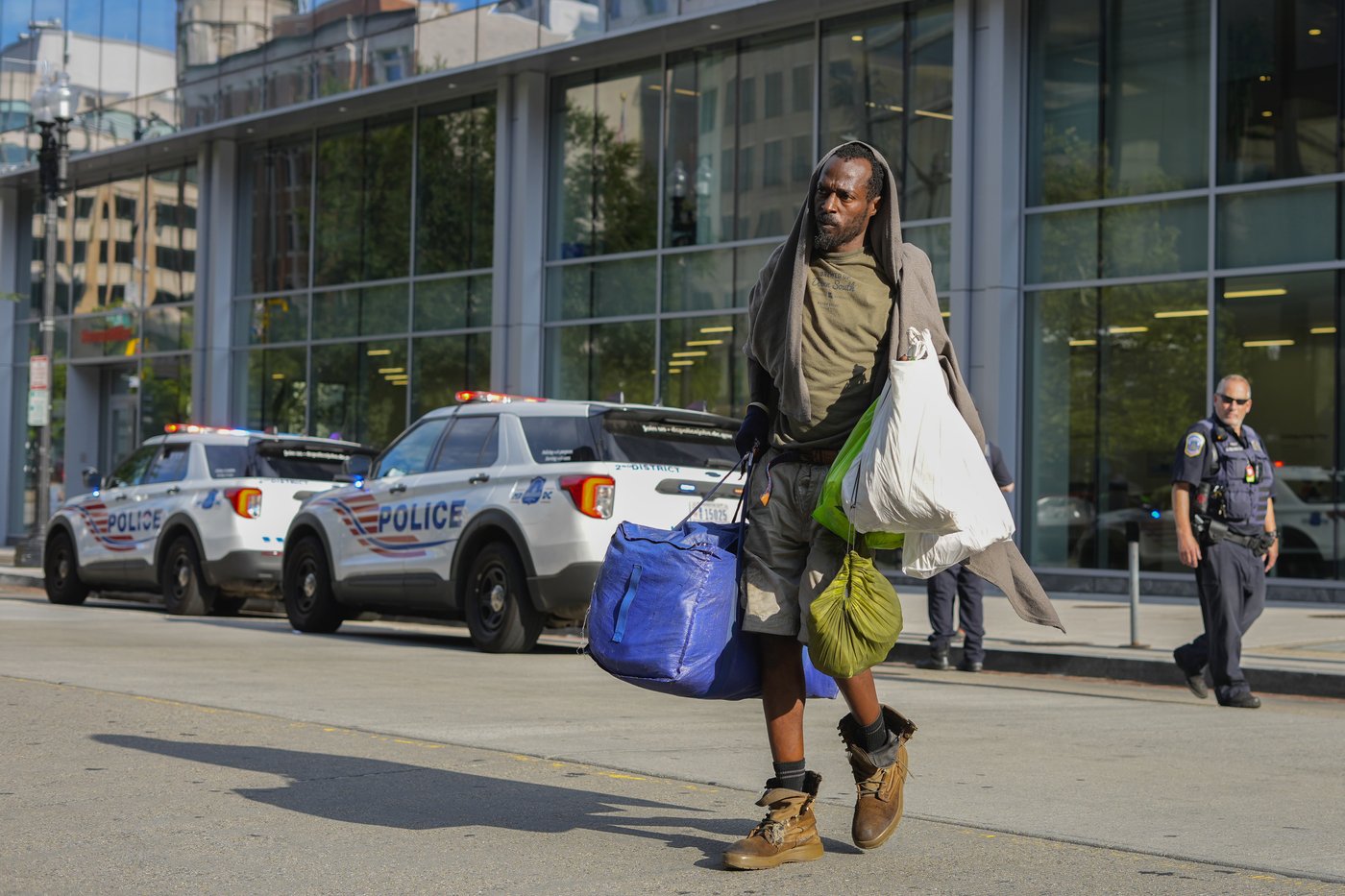 A man carring his personal belongings as he crosses the street infront of Washington Metropolitan Police officer outside of the Martin Luther King Memorial library in downtown Washington, Thursday, Wednesday, Aug. 13, 2025. (AP Photo/Pablo Martinez Monsivais)