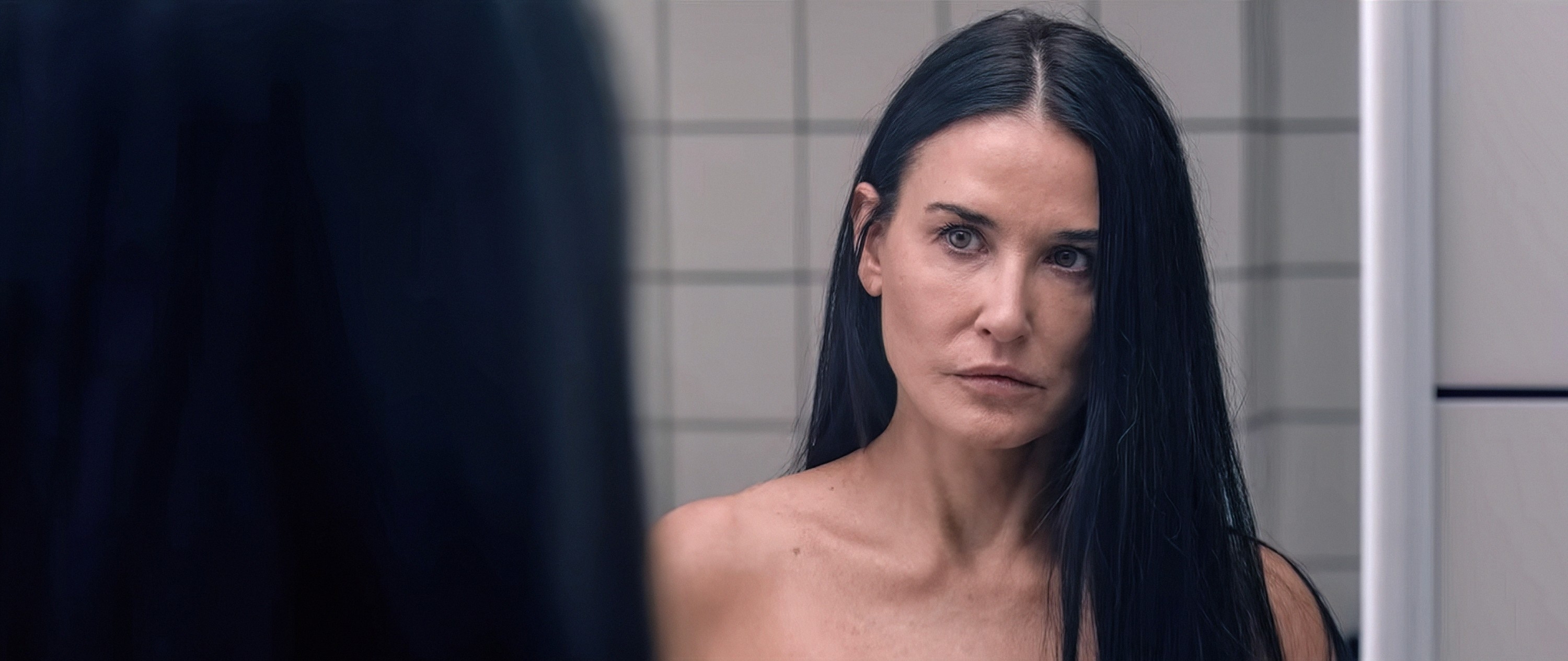 Person with long hair stares intently into a bathroom mirror, reflecting on something serious