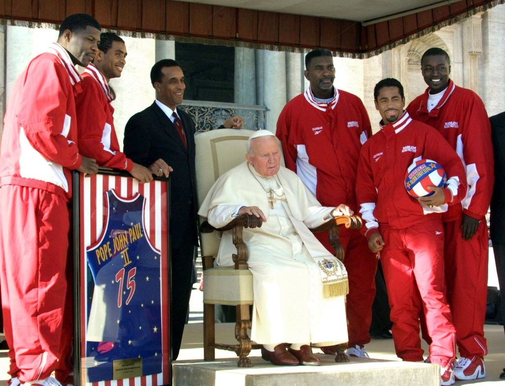 A group of basketball players in tracksuits posing with a religious leader seated in a chair, holding a basketball and a framed jersey