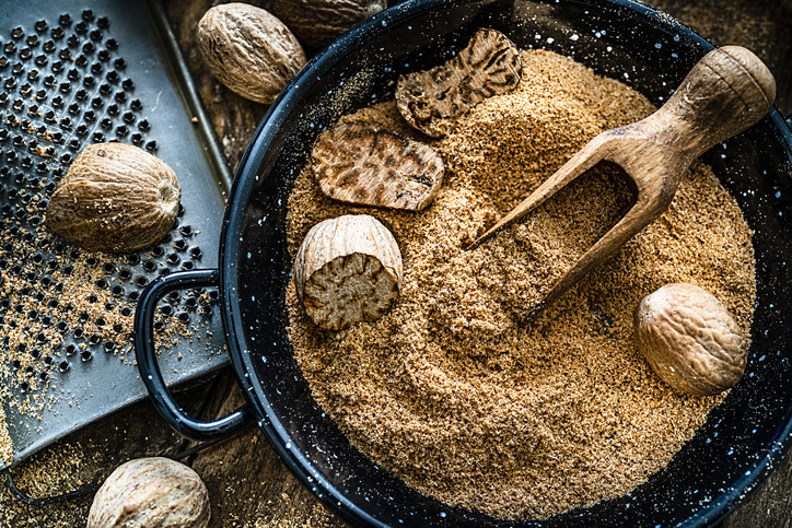 Nutmeg powder in a bowl with whole nutmeg seeds, a grater, and a wooden scoop. Rustic kitchen setting