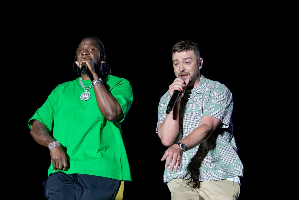 Two musicians perform on stage, one wearing a casual green t-shirt and the other a checkered short-sleeve shirt, both holding microphones
