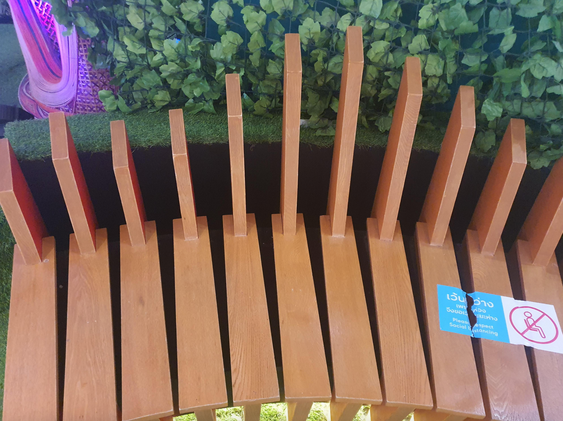 Wooden bench with uneven backrests, designed to deter lying down. Sign indicates sitting only, not for sleeping. Greenery in the background