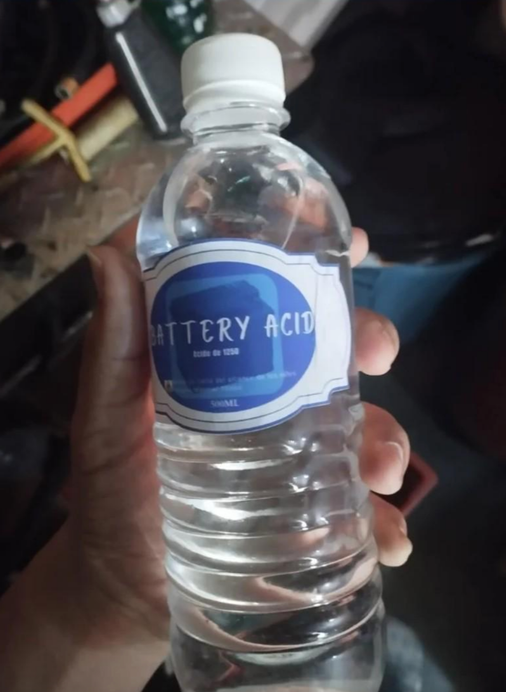 A hand holds a water bottle labeled "Battery Acid" in a casual setting