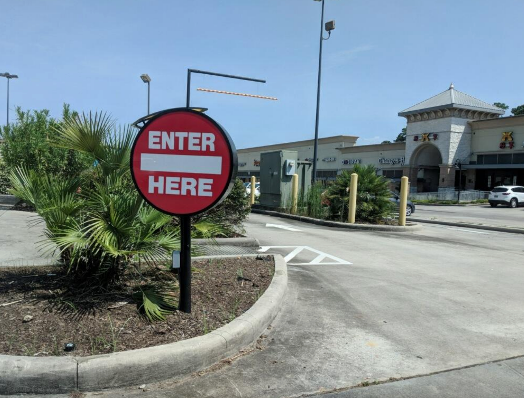 A large round sign reads "ENTER HERE" in front of a shopping plaza, directing cars into a parking area