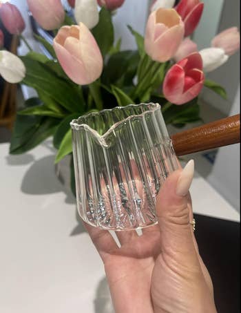 Hand holding a small glass coffee dripper with a wooden handle; tulips are in the background