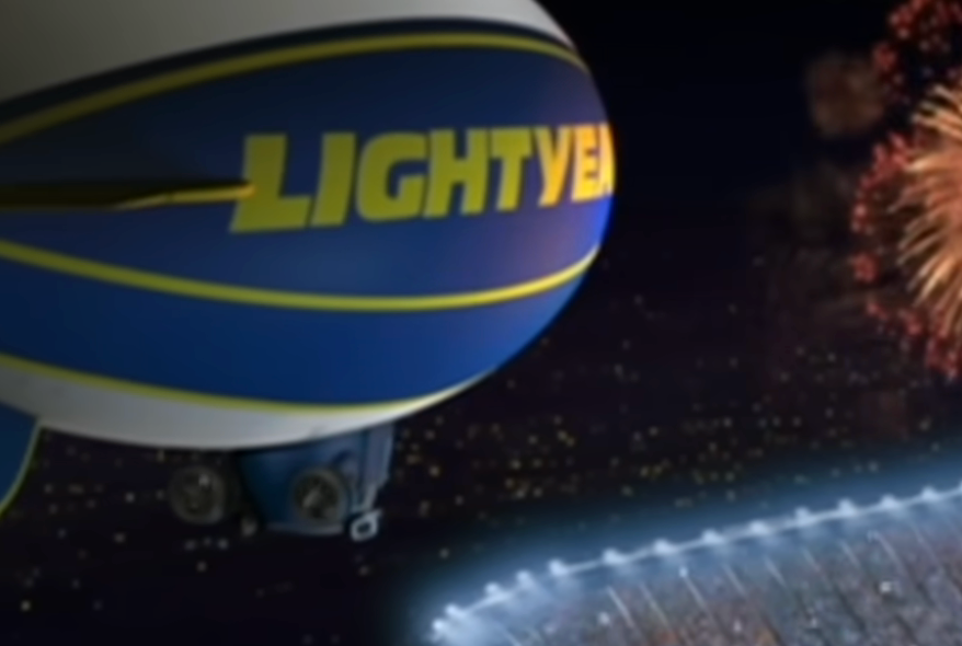 A blimp labeled "Lightyear" is flying near a stadium at night, with fireworks in the background