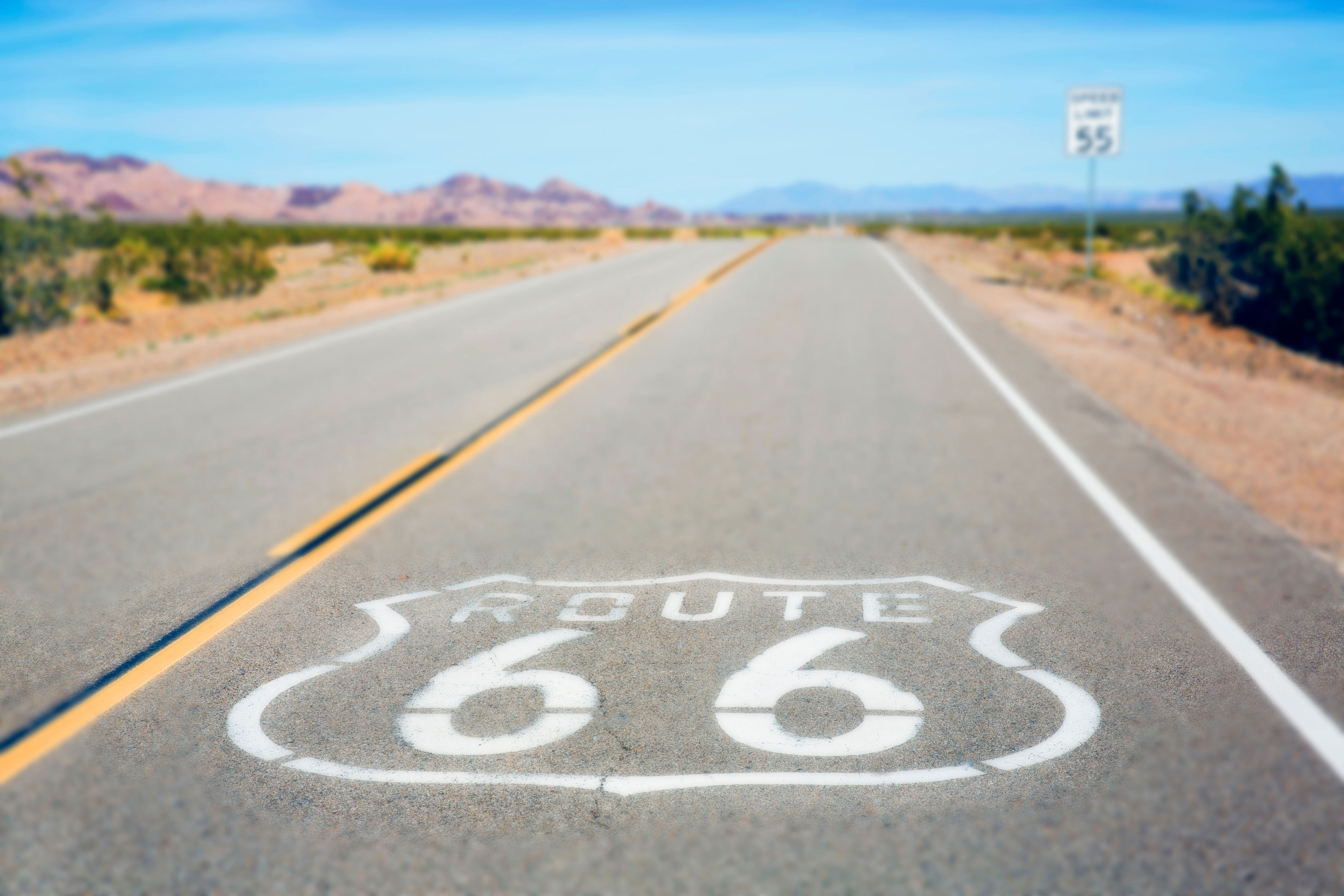 Open highway with "Route 66" emblem painted on the road, stretching into a desert landscape. Speed limit sign visible in the distance