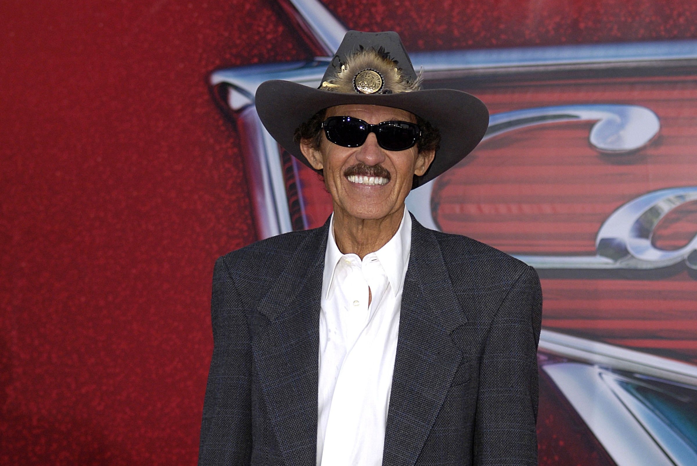 A person in a cowboy hat and sunglasses wears a plaid blazer, white shirt, and jeans on a red carpet at a movie premiere