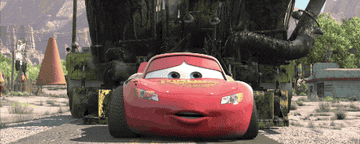 Animated car with big eyes and a smiling mouth speeds in front of a large asphalt truck on a desert road, from a movie scene