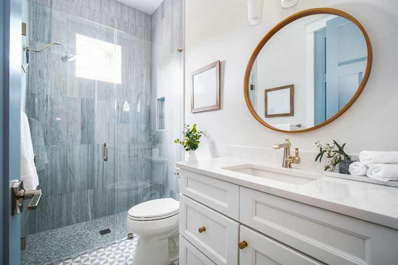 Modern bathroom featuring a glass shower, white vanity with gold fixtures, round mirror, and decorative plants.