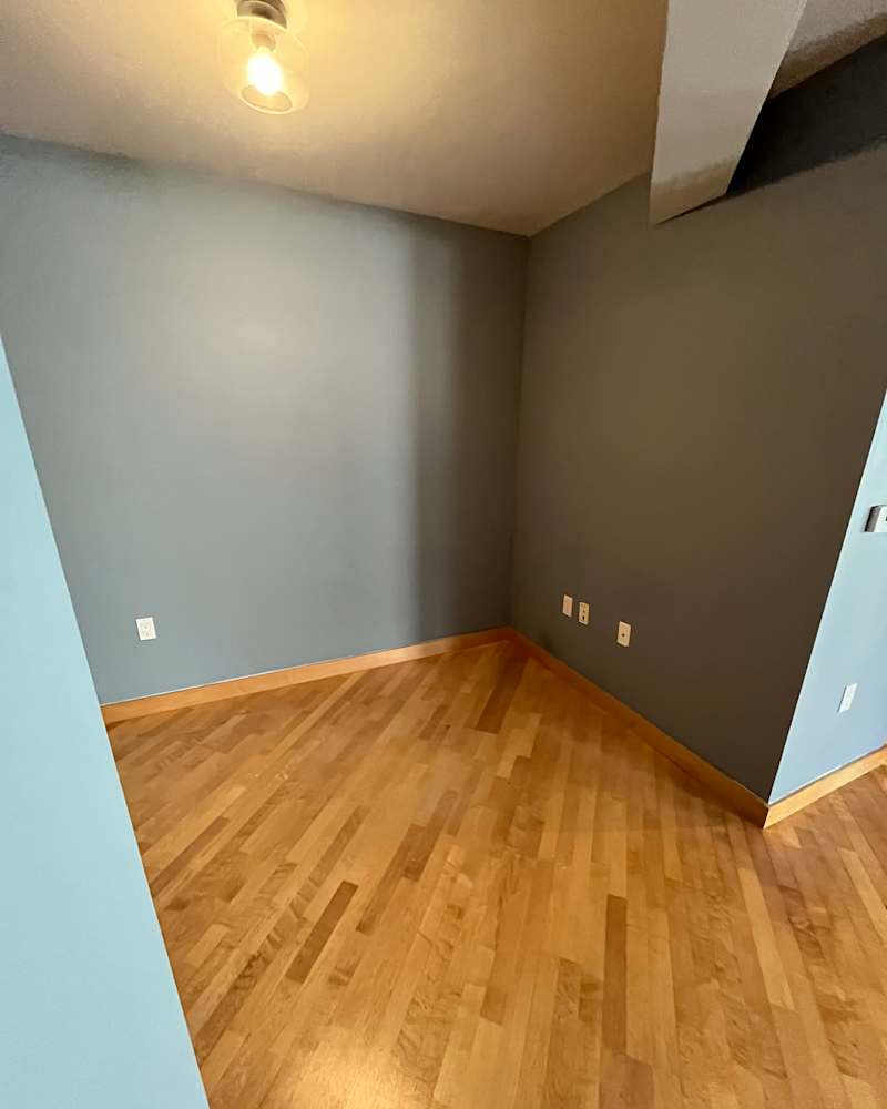 Empty corner of a room with light wood flooring, gray walls, and a ceiling light fixture.