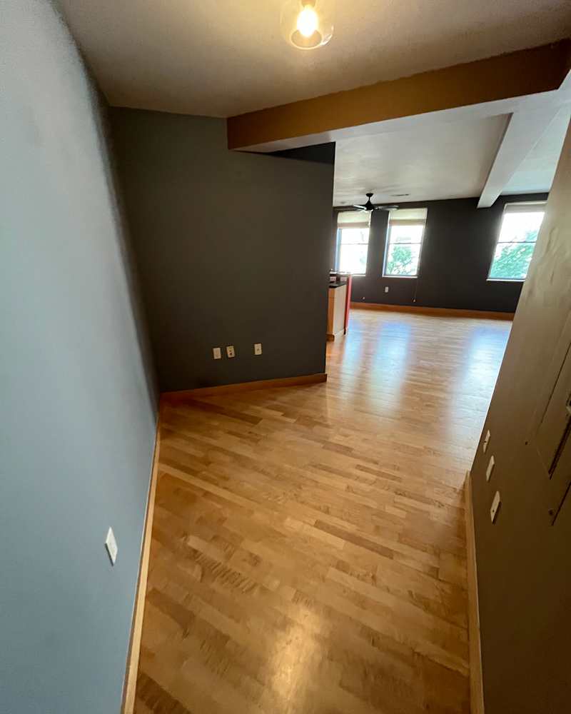 Spacious room with hardwood flooring, gray walls, and natural light from windows, leading to an open area.