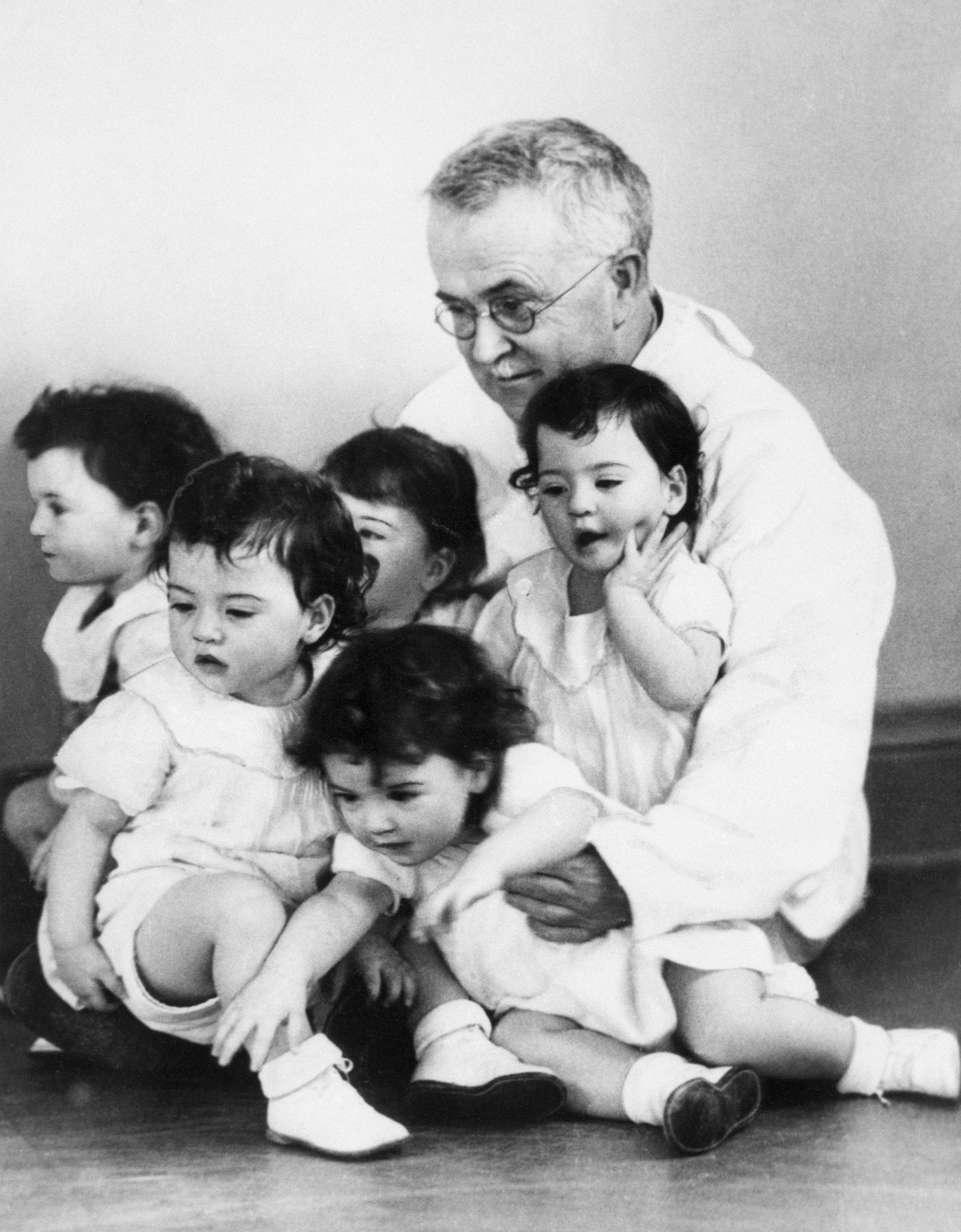 [Original caption] Honoured guest Dr. Allan R. Dafoe puts his arms around all five of the Dionne quintuplets Marie, Emelie, Cecile, Annette and Yvonne as they celebrate their first birthday in style