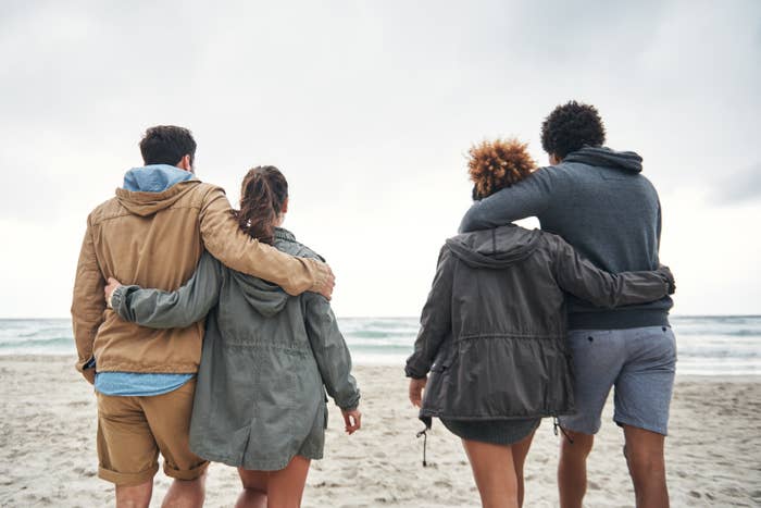 Two couples walk arm-in-arm along a beach, dressed casually in jackets and shorts, emphasizing companionship and affection