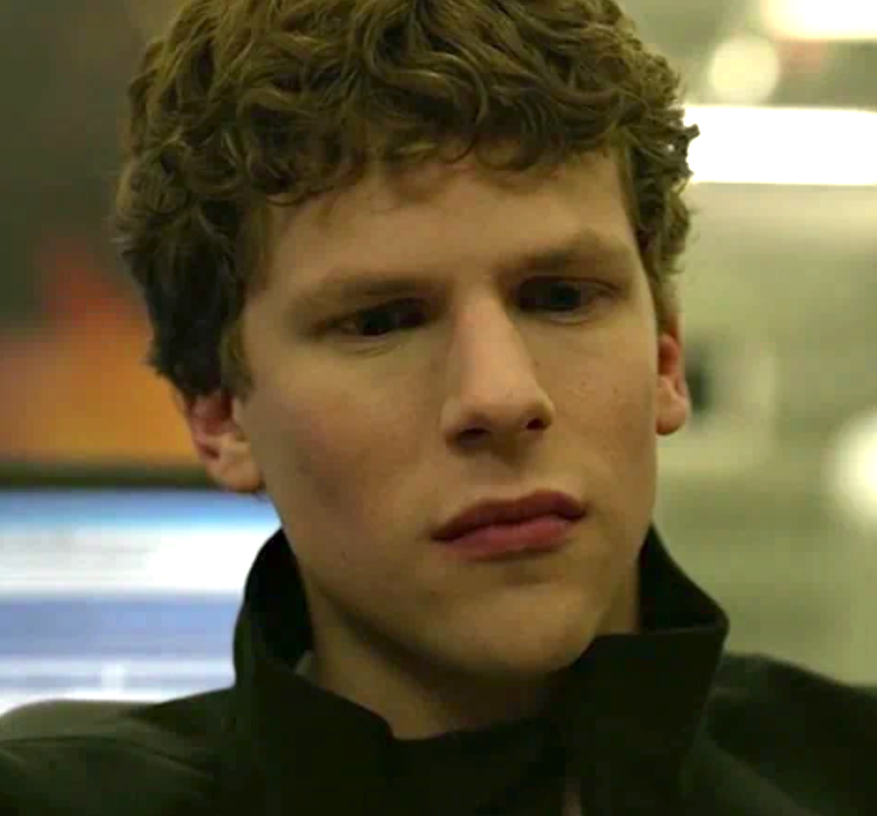 A person with curly hair looks thoughtful, wearing a jacket with a popped collar, appearing to be in a technology-focused setting
