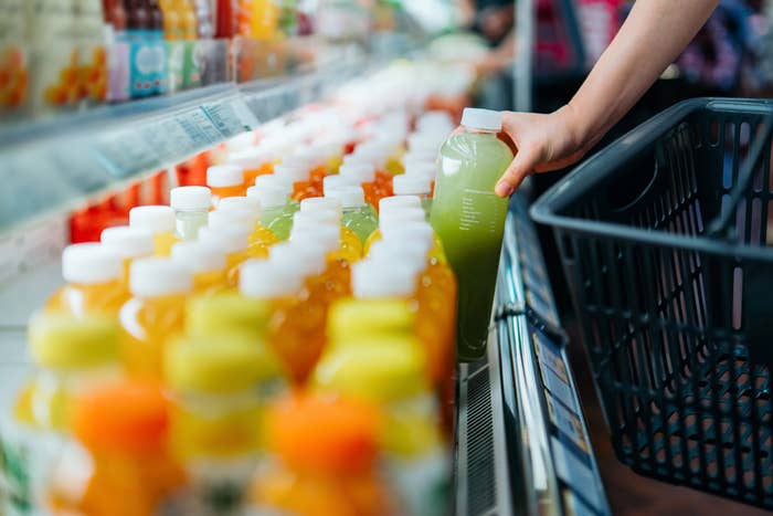 Person selects a green juice bottle from a supermarket shelf filled with various beverages, holding a shopping basket in their other hand