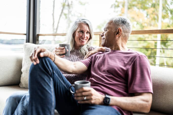 Older couple sits on a couch with mugs, smiling at each other, creating a warm, intimate moment