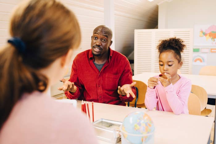 A man and a child sit at a table in a classroom setting, in discussion with a woman whose back is to the camera. The child focuses on an activity