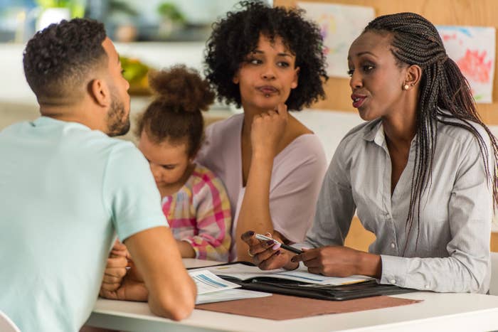 Two parents talk with a teacher at a table during a parent-teacher meeting. A young child sits quietly, drawing with crayons