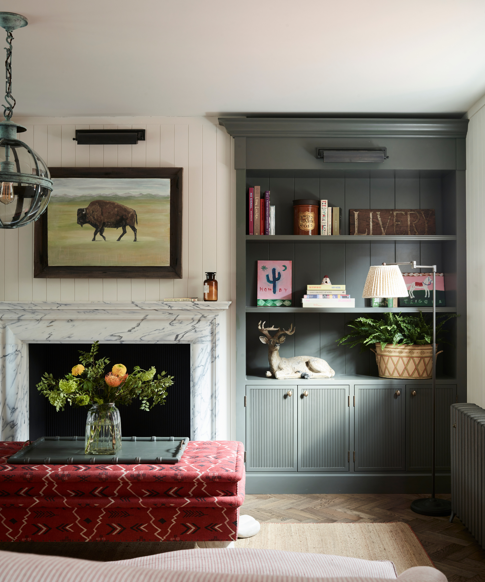 a living room with a marble fireplace, a photo of a buffalo on the wall, a built in shelving unit with books and decor, and a red printed ottoman with a tray and flowers on top