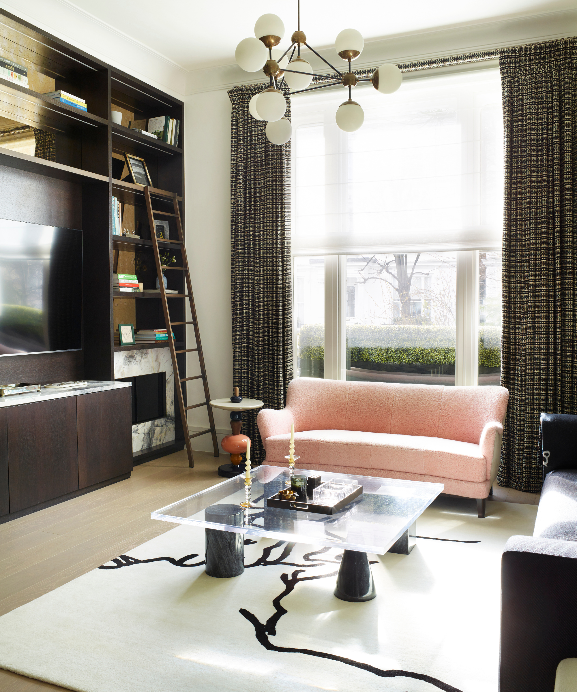 a living room with a large built-in shelving unit, tall windows with dark brown curtains, a pink sofa, a glass coffee table, and a white rug with black detailing