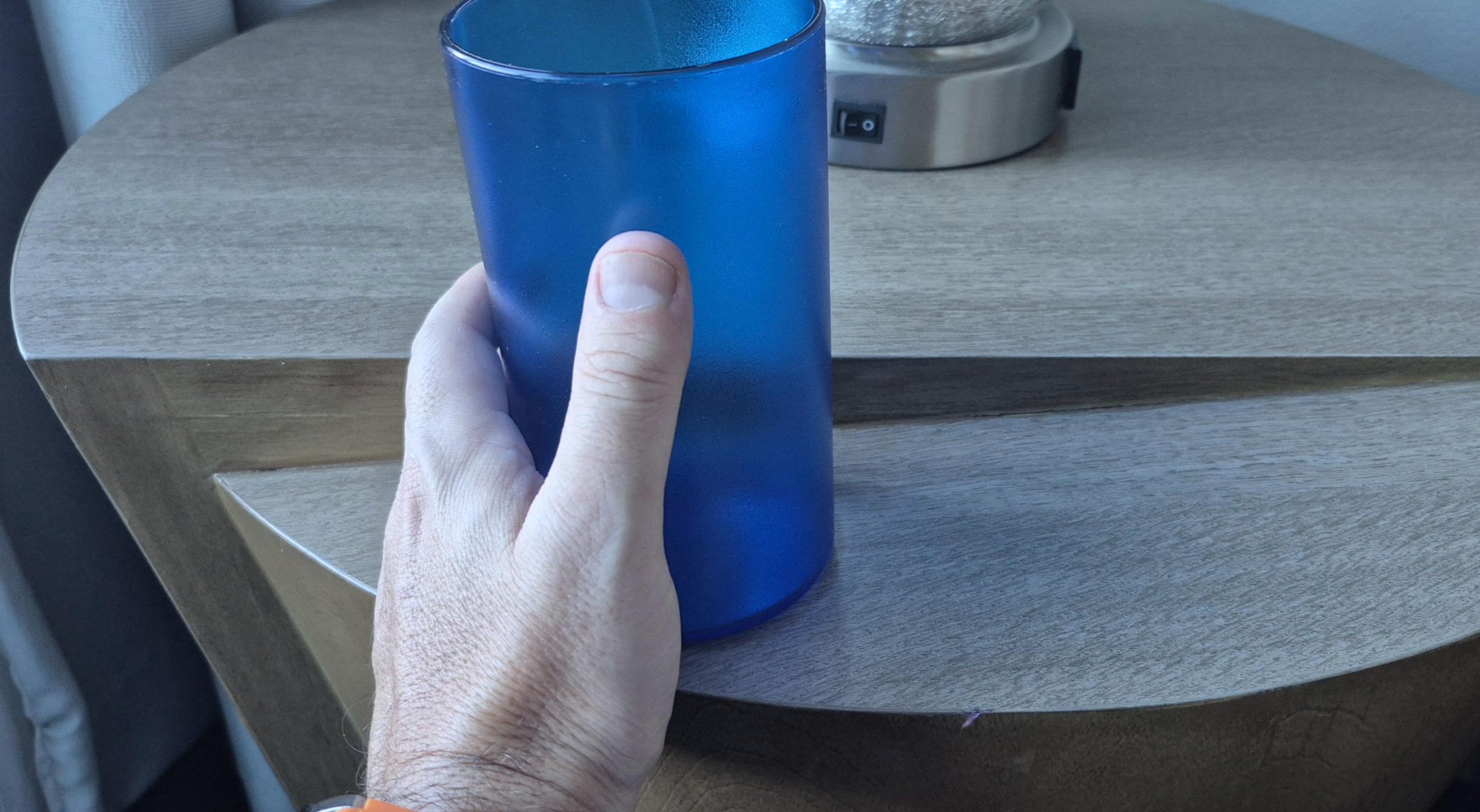 A hand holding a blue glass in front of a round, wooden table with a small, cylindrical lamp