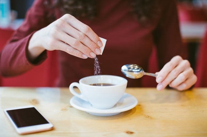 Woman’s hands pouring sugar into black coffee – girl sitting at the table with espresso and smartphone – blood and glycemic index control for diabetes -excess of white sugar in food concept