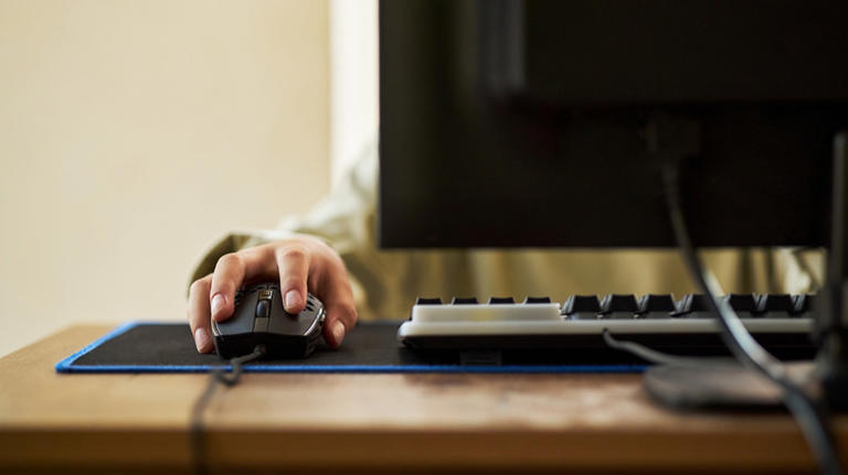 A person using a keyboard and mouse with an out of focus monitor in the foreground