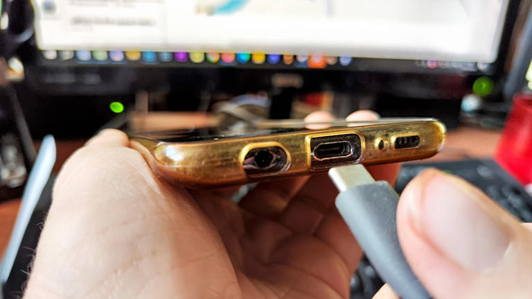 A person plugging their smartphone to charge with a monitor in the background