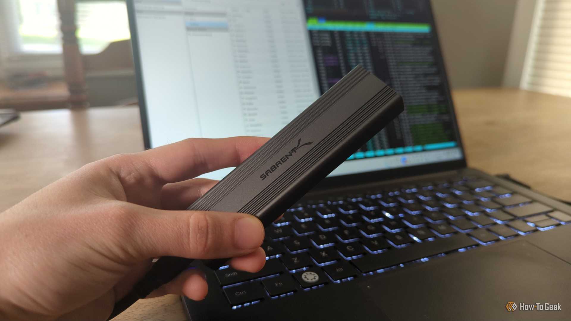 A person's hand holding a Sabrent external SSD drive in front of a Linux laptop.