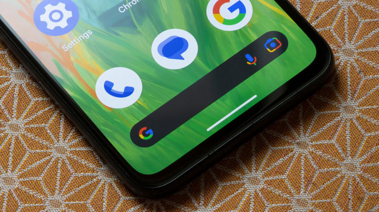 An Android smartphone laying on a patterned table cloth displaying the Home screen, emphasizing the Google Search bar widget