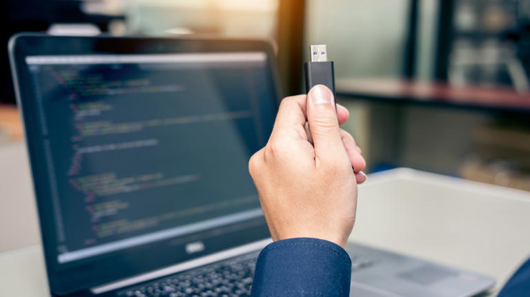 A hand holding a USB flash drive with a laptop in the background