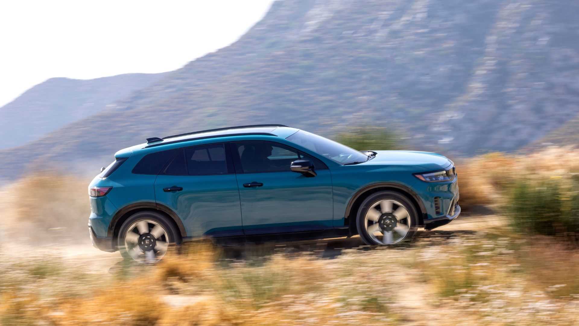 Dynamic side profile shot of a blue 2025 Honda Prologue driving on a dirt track with mountains in the background.