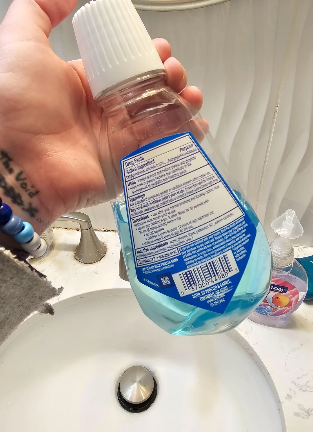 A hand holds a bottle of mouthwash over a bathroom sink, showing the back label detailing active ingredients and usage instructions