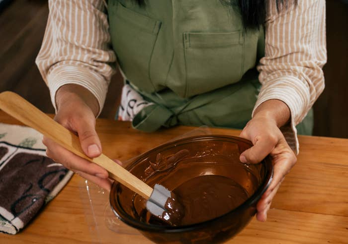 Person holding a bowl of melted chocolate with a spatula, next to a bowl of chocolate pieces on a wooden table