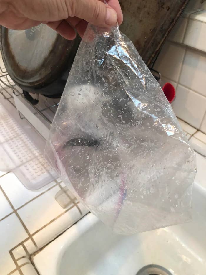 A hand holding an empty, wet plastic bag above a kitchen sink with drying dishes in the background