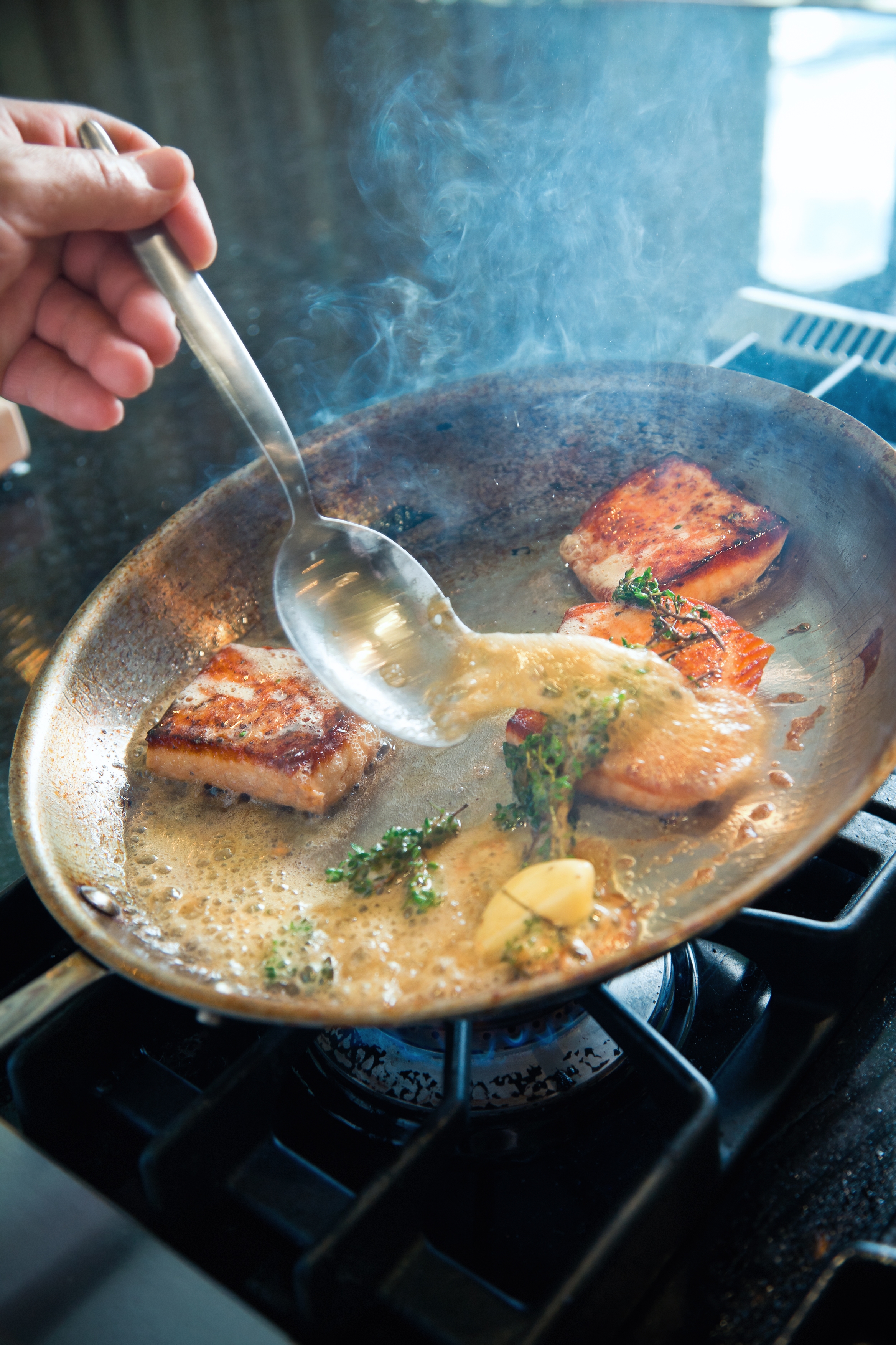 Seared salmon fillets are being basted with butter sauce in a pan on a stovetop