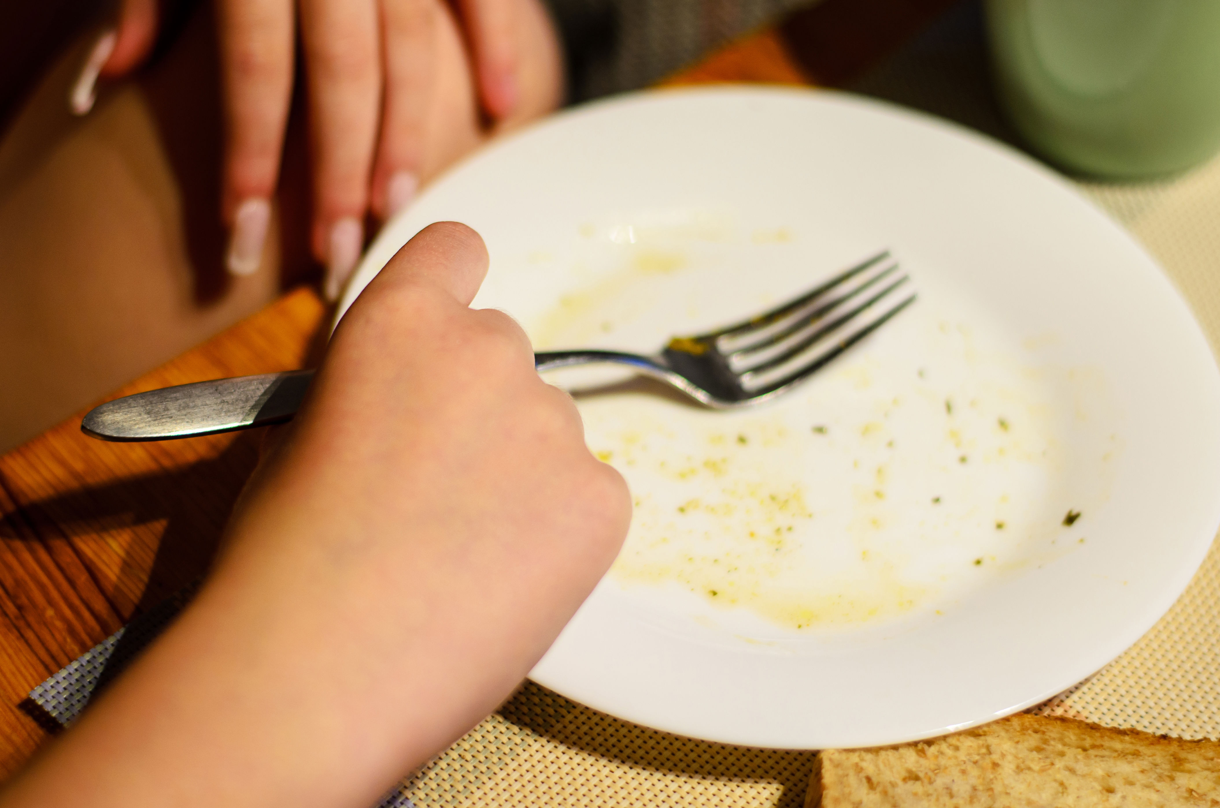 A hand holds a fork over an empty plate with food crumbs, suggesting completion of a meal, on a table with another hand resting nearby