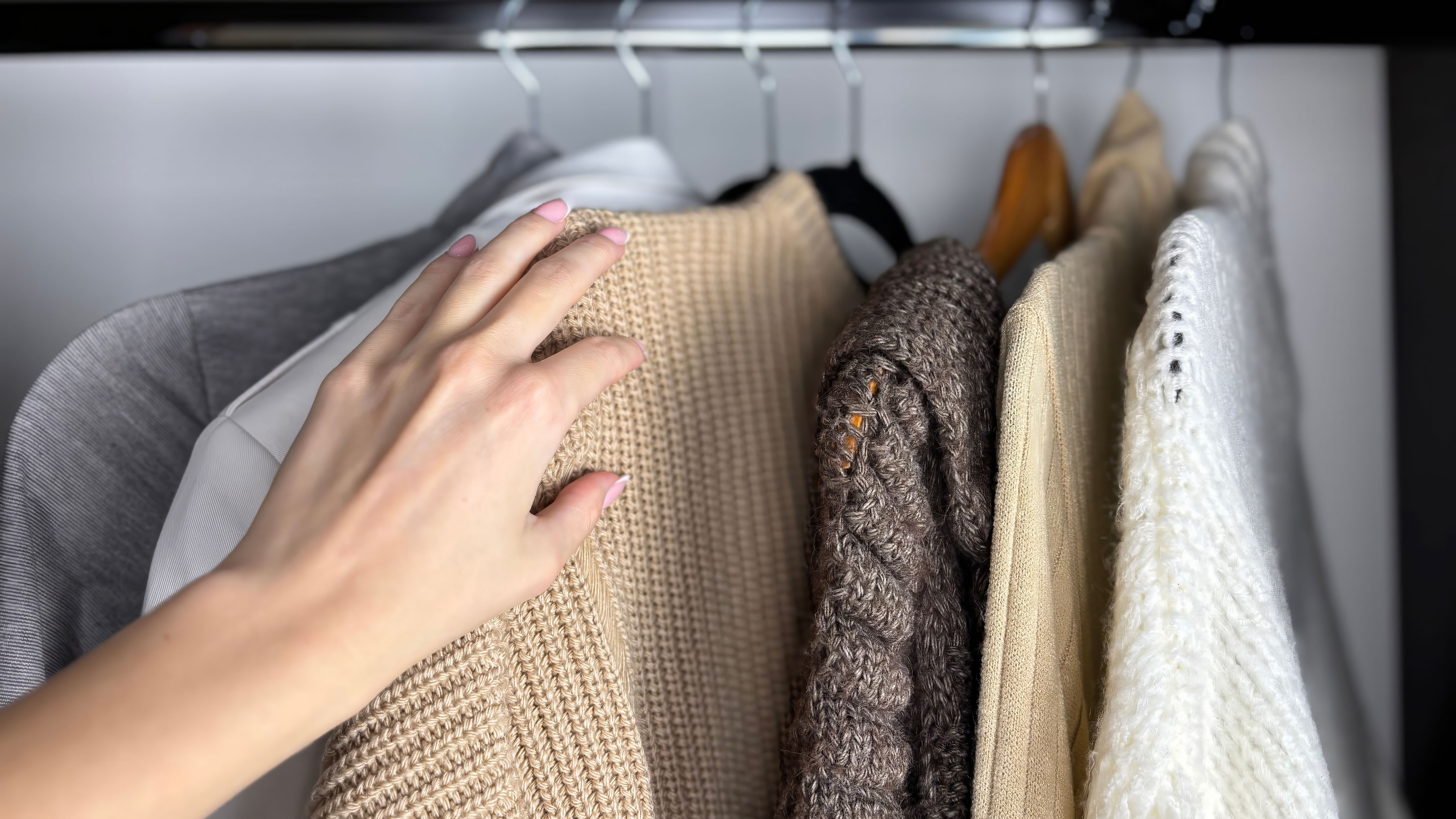 A hand touches various knit sweaters hanging in a closet