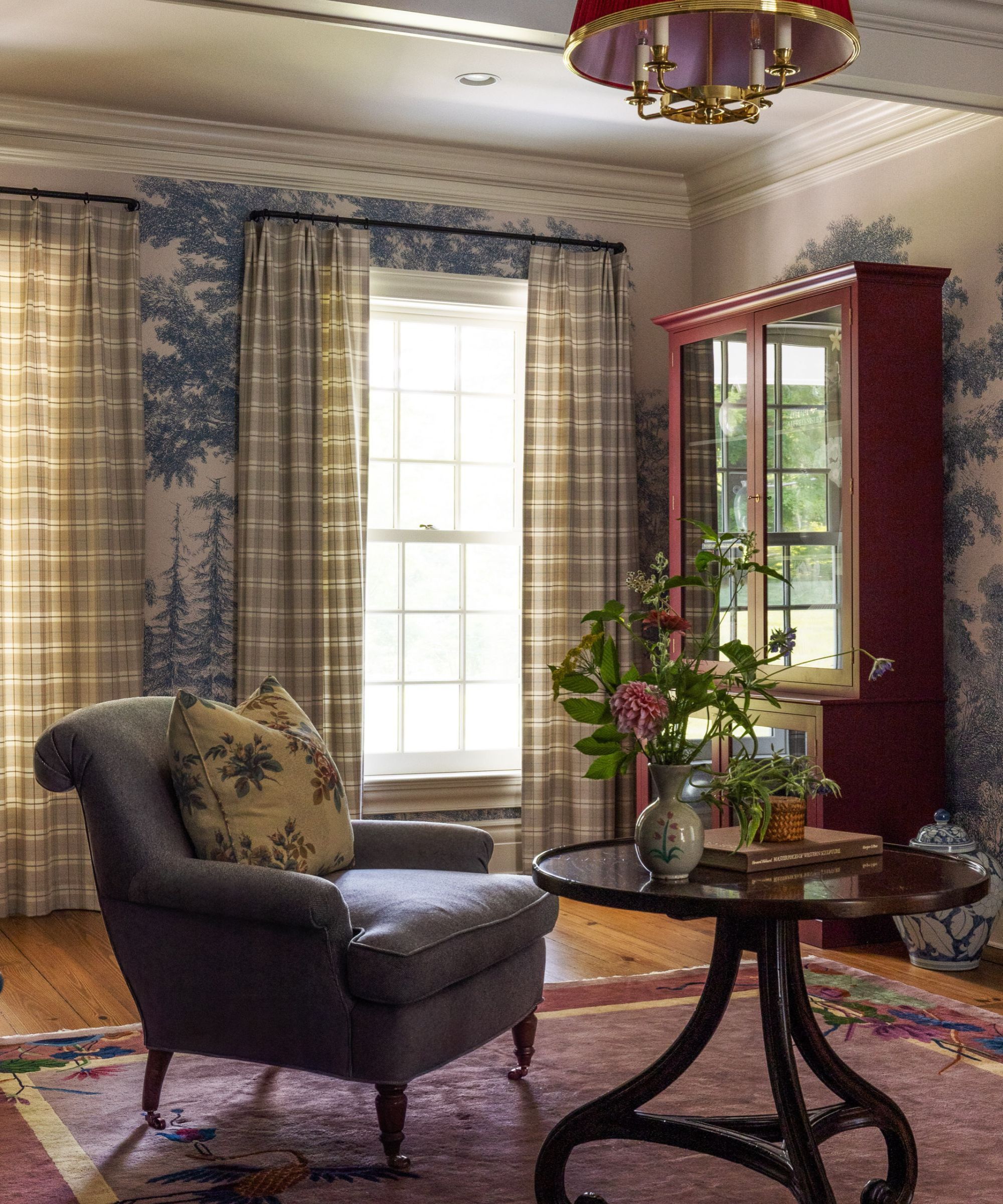 cozy living room armchair and table with blue and white tree mural wallpaper, gingham drapes, wooden dresser and flowers in a vase