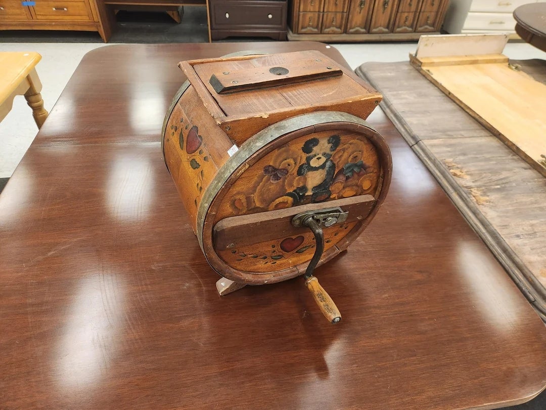 Vintage hand-cranked drum grater with floral design on side, sitting on a wooden table among various furniture items