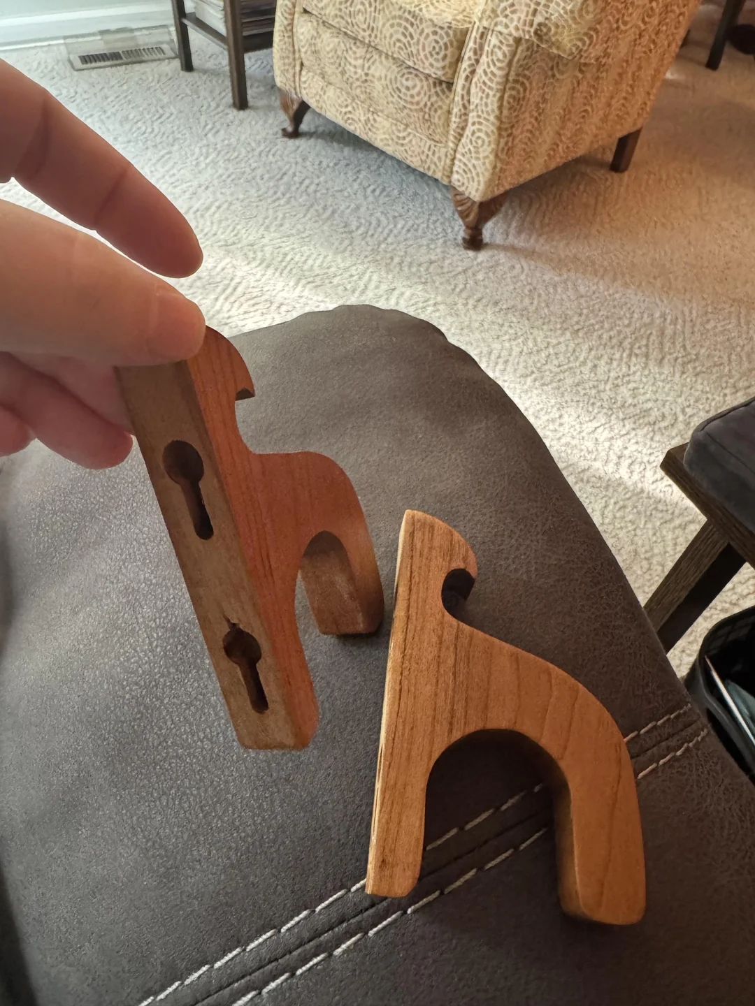 Hand holding a wooden tool with keyhole shapes; another similar tool rests on a couch. The setting is a living room