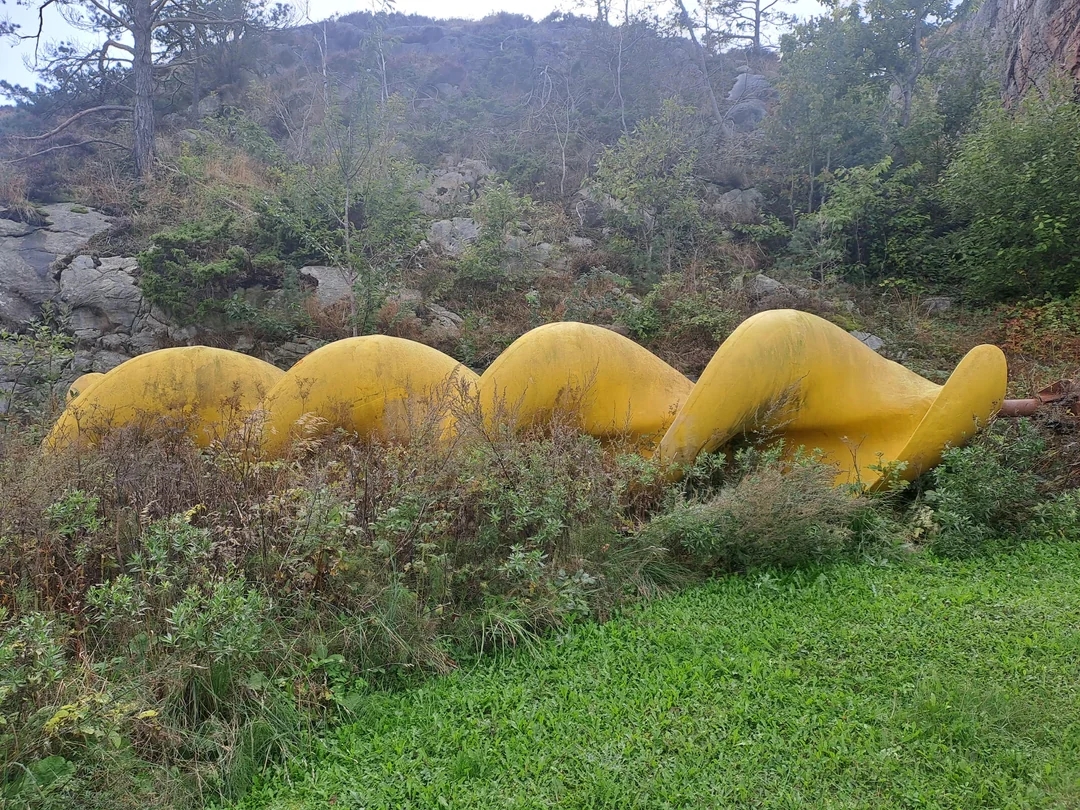 Large yellow spiral structure partially hidden in grassy, wooded area with rocks. It appears abandoned or part of an outdoor installation